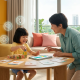 Child learning with parent in a sunlit Singapore apartment, colorful materials, AI motifs.